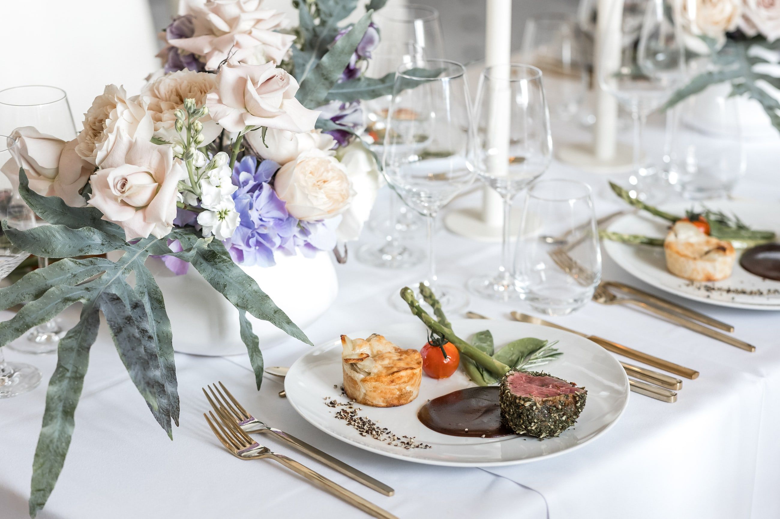 Festlich eingedeckter Tisch mit Rinderfilet, Gratin und Gemüse auf weißem Porzellan, daneben Kristallgläser und ein Blumengesteck