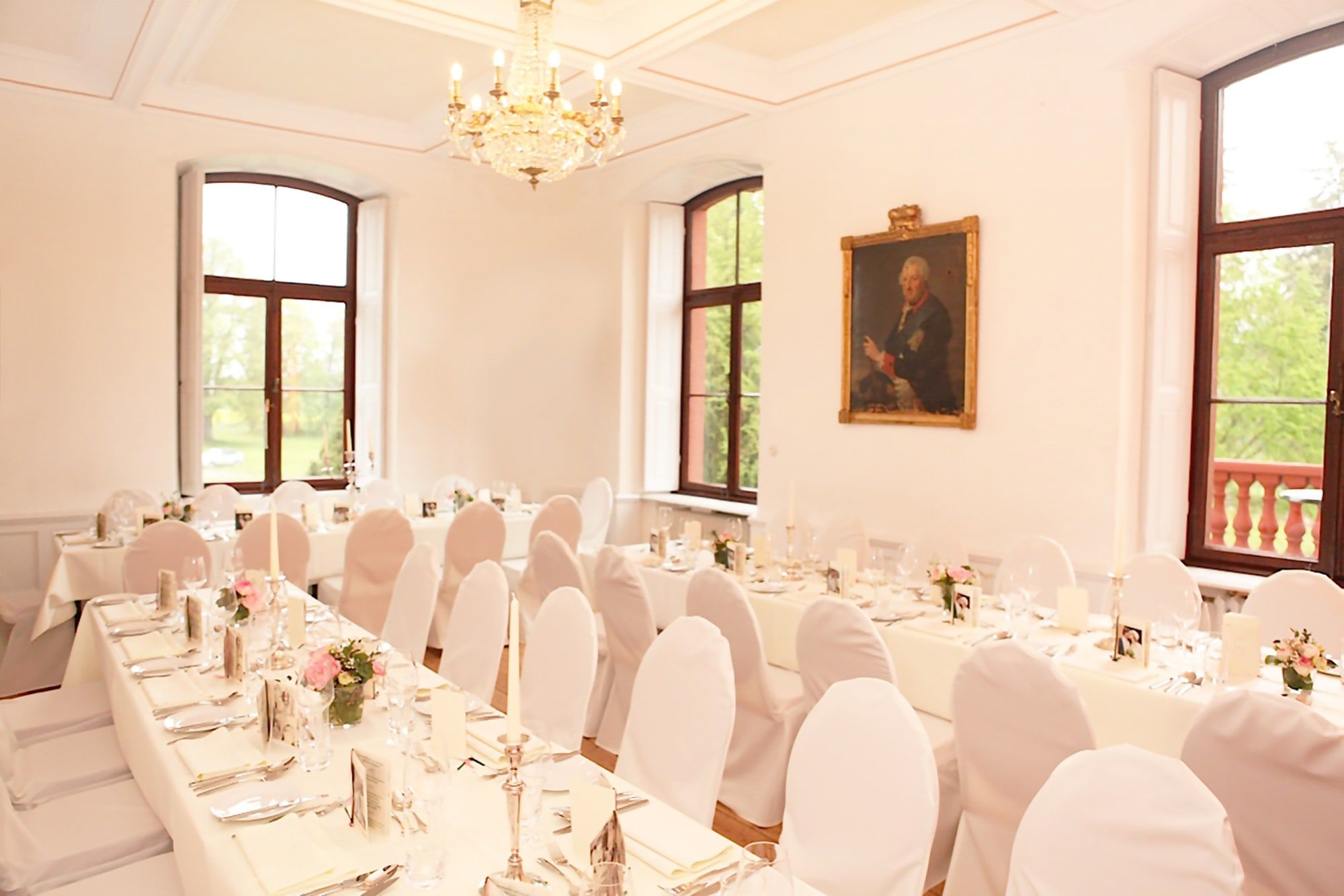 Heller Festsaal auf Schloss Wachendorf mit Kronleuchter, Ahnenporträt und weiß eingedeckten Tafeln vor hohen Fenstern zum Park