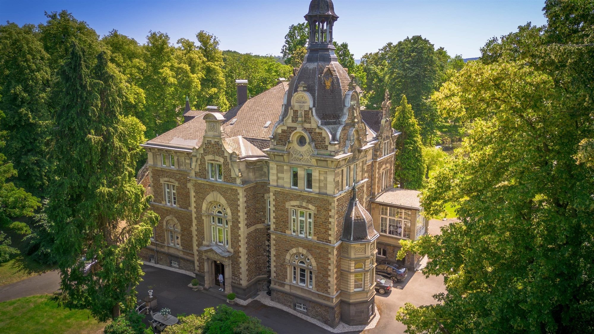 Luftaufnahme von Schloss Vettelhoven mit Turmhaube, Sandsteinfassade und den hohen Bäumen des Parks