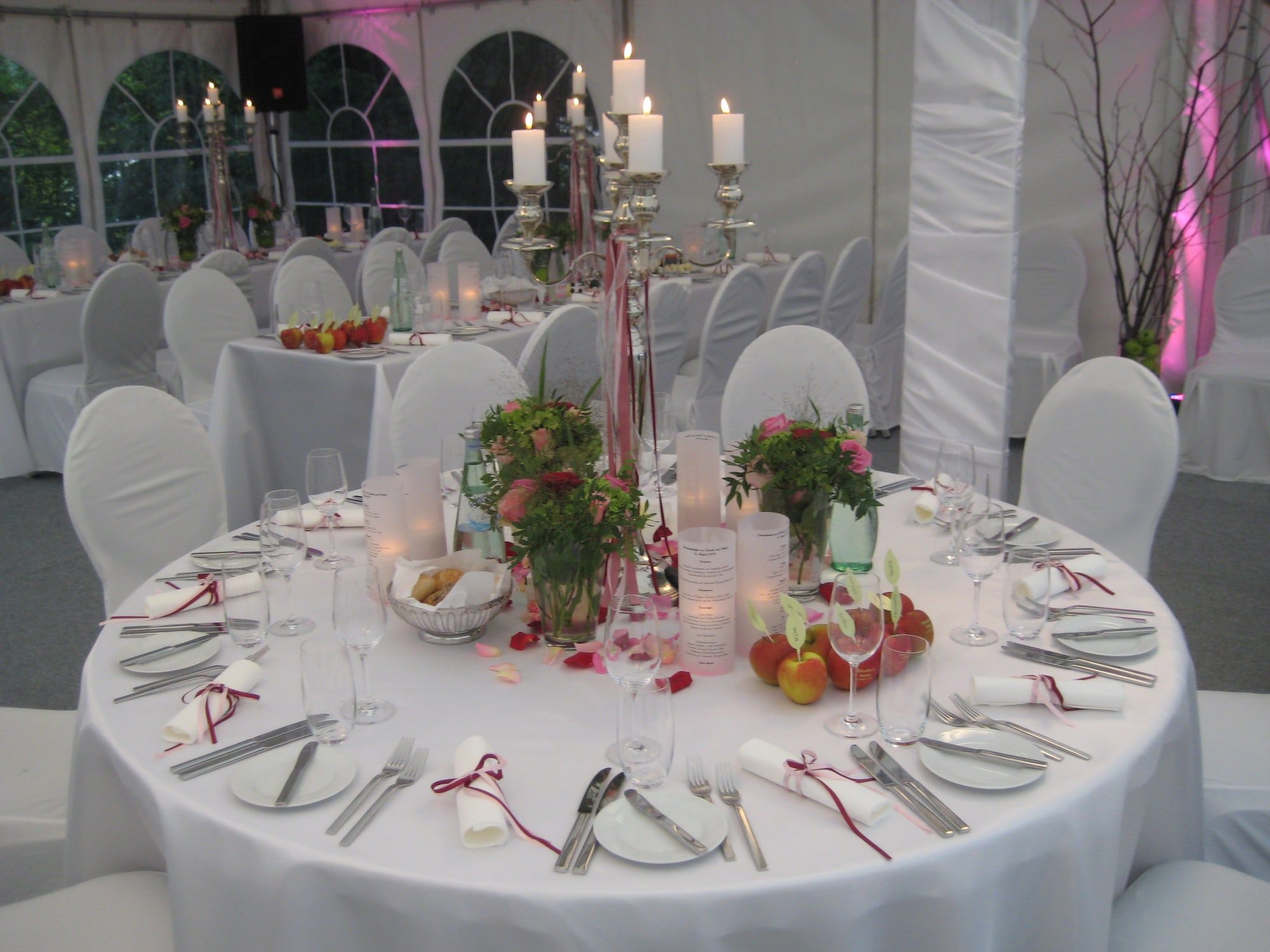 Rund gedeckte Hochzeitstafel im Festzelt mit silbernem Kerzenleuchter, Rosen, Äpfeln und weißen Hussen, dahinter Rundbogenfenster