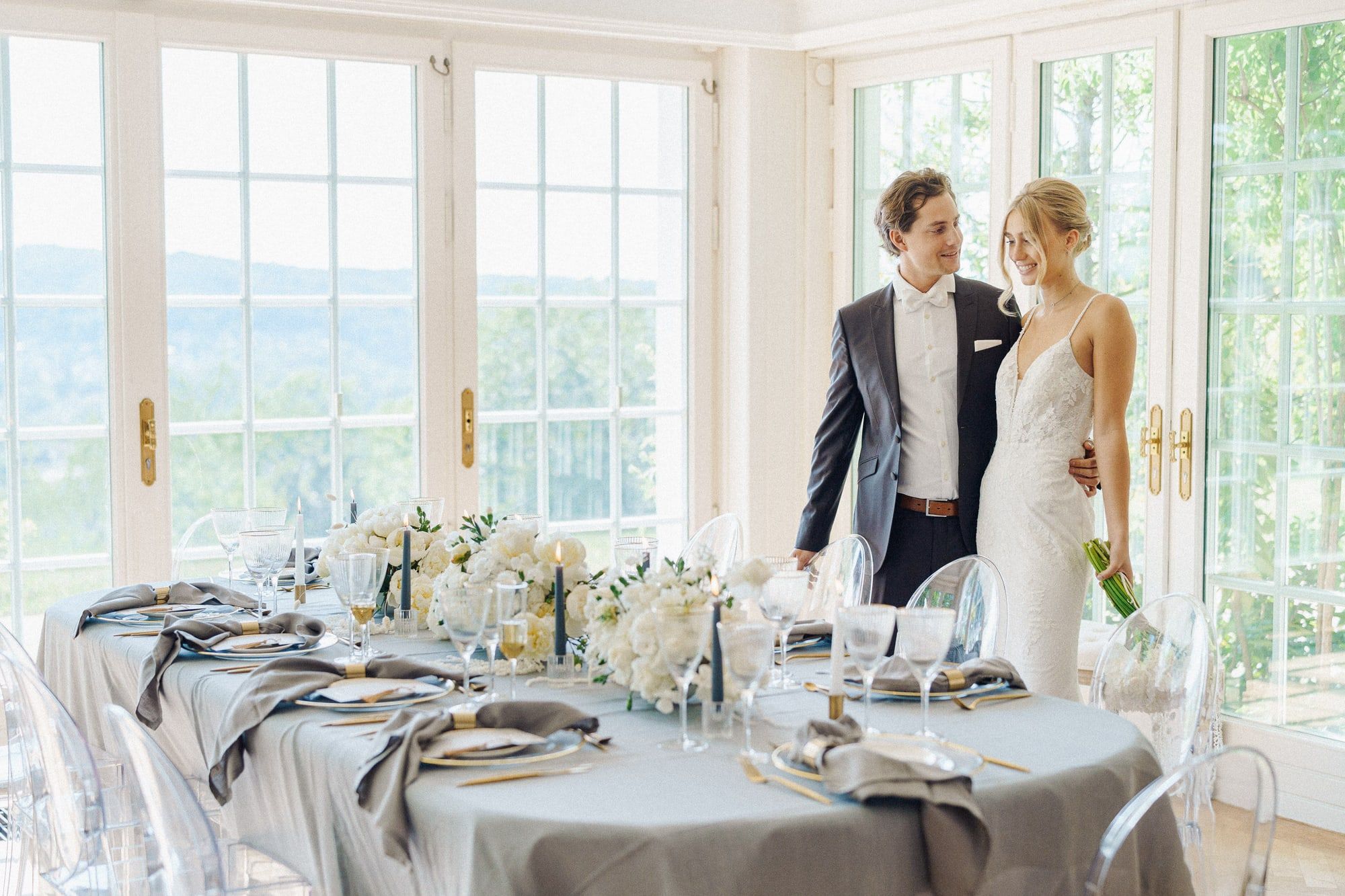 Brautpaar an der eingedeckten Hochzeitstafel vor den hohen Sprossenfenstern der Villa Heckenfels mit Blick ins Grüne