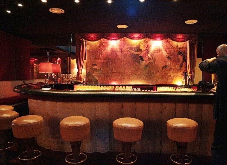 Geschwungene Theke der Kokett Bar mit cremefarbenen Barhockern vor einem großen Wandgemälde im Stil der Belle Époque