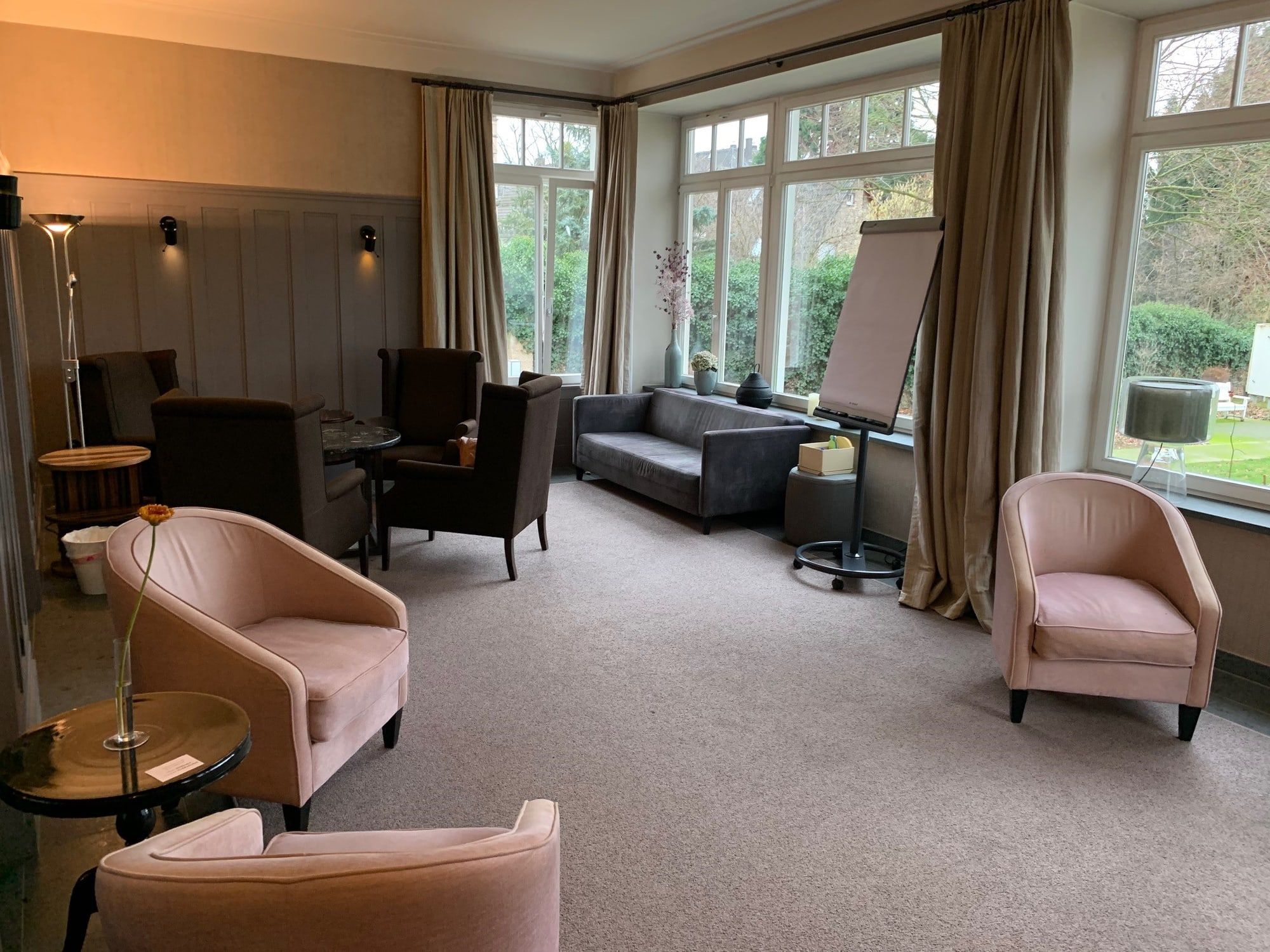 Wintergarten-Lounge der Villa Leonhart mit Sesseln, Sofa und großen Fenstern, durch die man ins Grün des Parks blickt