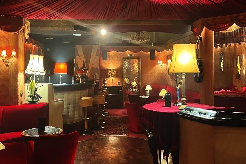 Barraum der Kokett Bar in Köln mit Lampenschirmen, Kristallleuchter, Spiegeltür und roten Samtmöbeln im warmen Licht