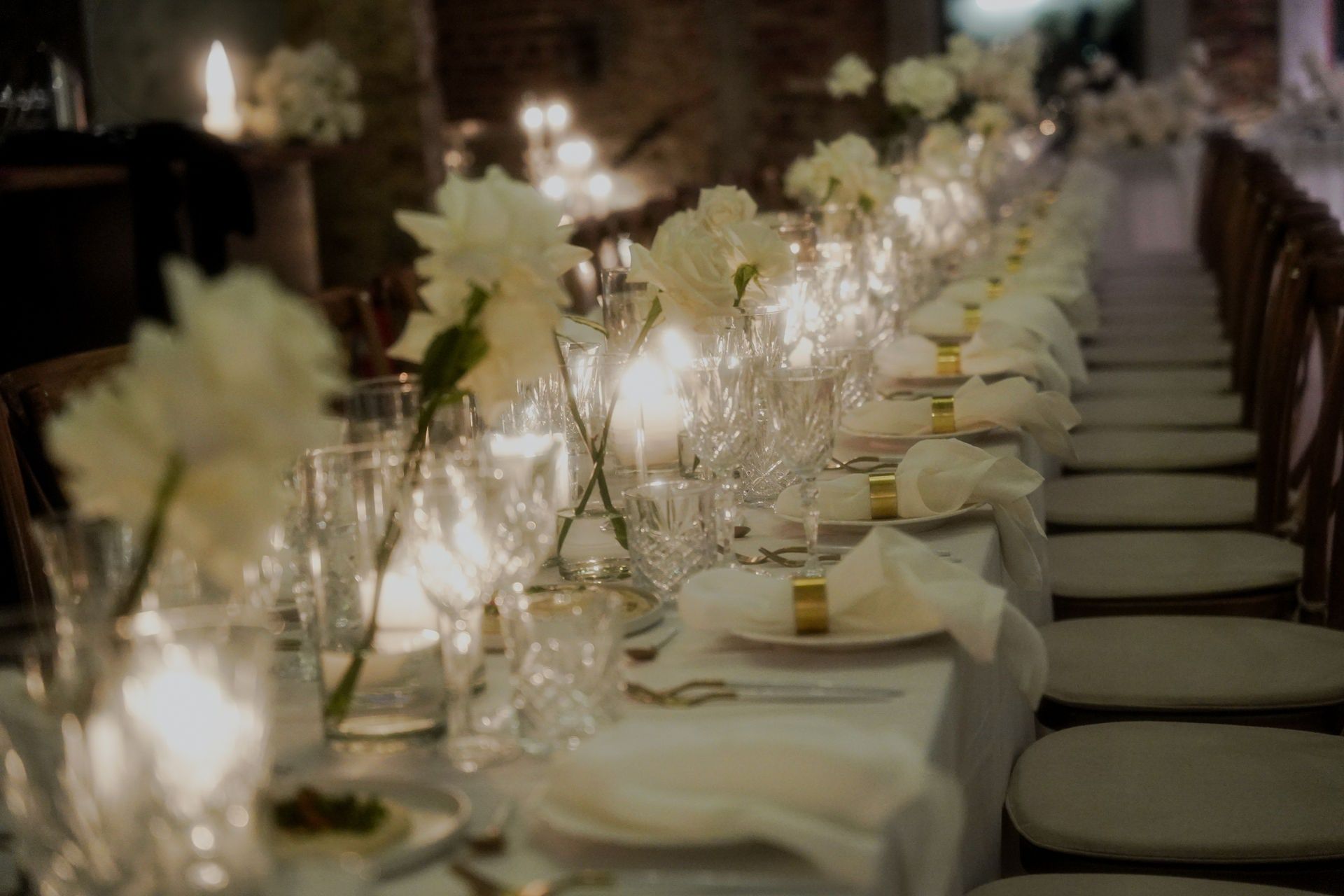 Lange Hochzeitstafel im Kerzenlicht mit weißen Blumen, Kristallgläsern und goldenen Serviettenringen im Andreashaus