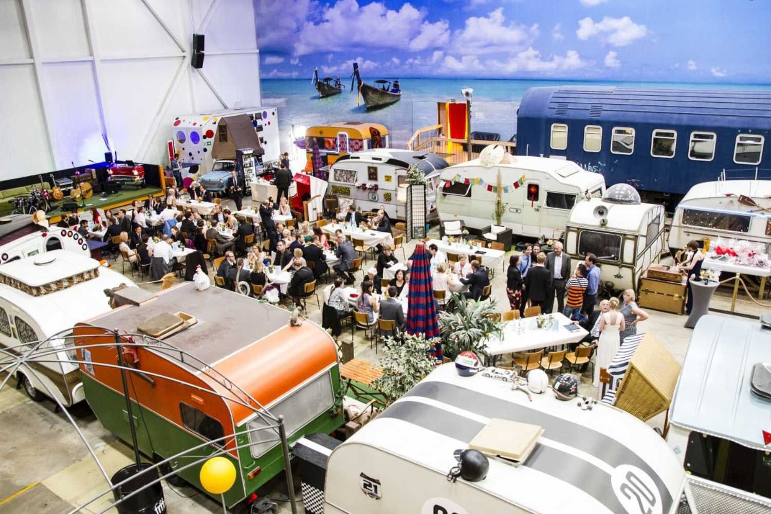 Hochzeitsgesellschaft beim Essen an Tischen zwischen bunten Oldtimer-Wohnwagen, im Hintergrund ein Schlafwagen und ein gemalter Strand