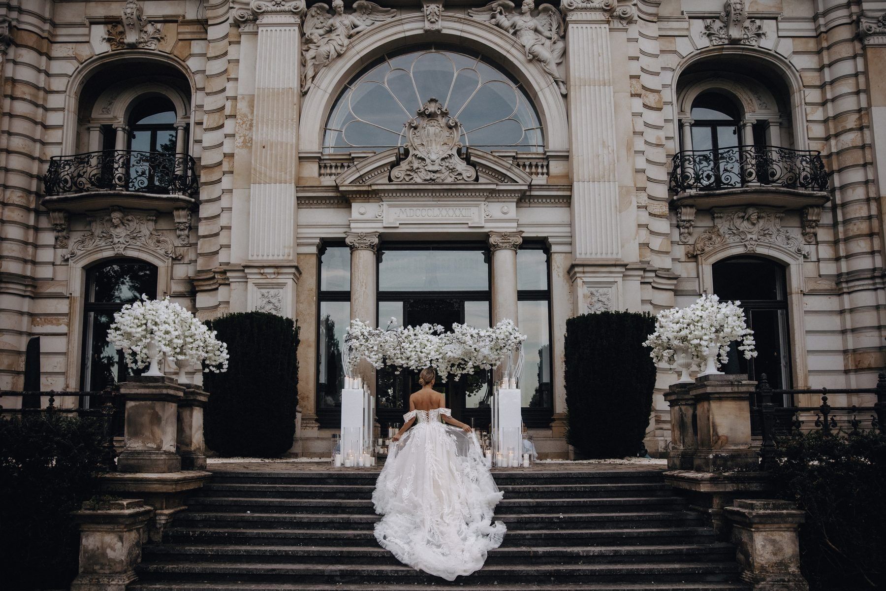Eine Braut im ausladenden Kleid schreitet die Freitreppe hinauf zum blumengeschmückten Portal der Villa