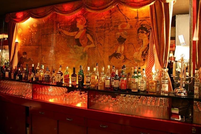 Beleuchtetes Rückbuffet der Bar mit Gläsern und Flaschen vor einem gemalten Varieté-Motiv und roten Vorhängen