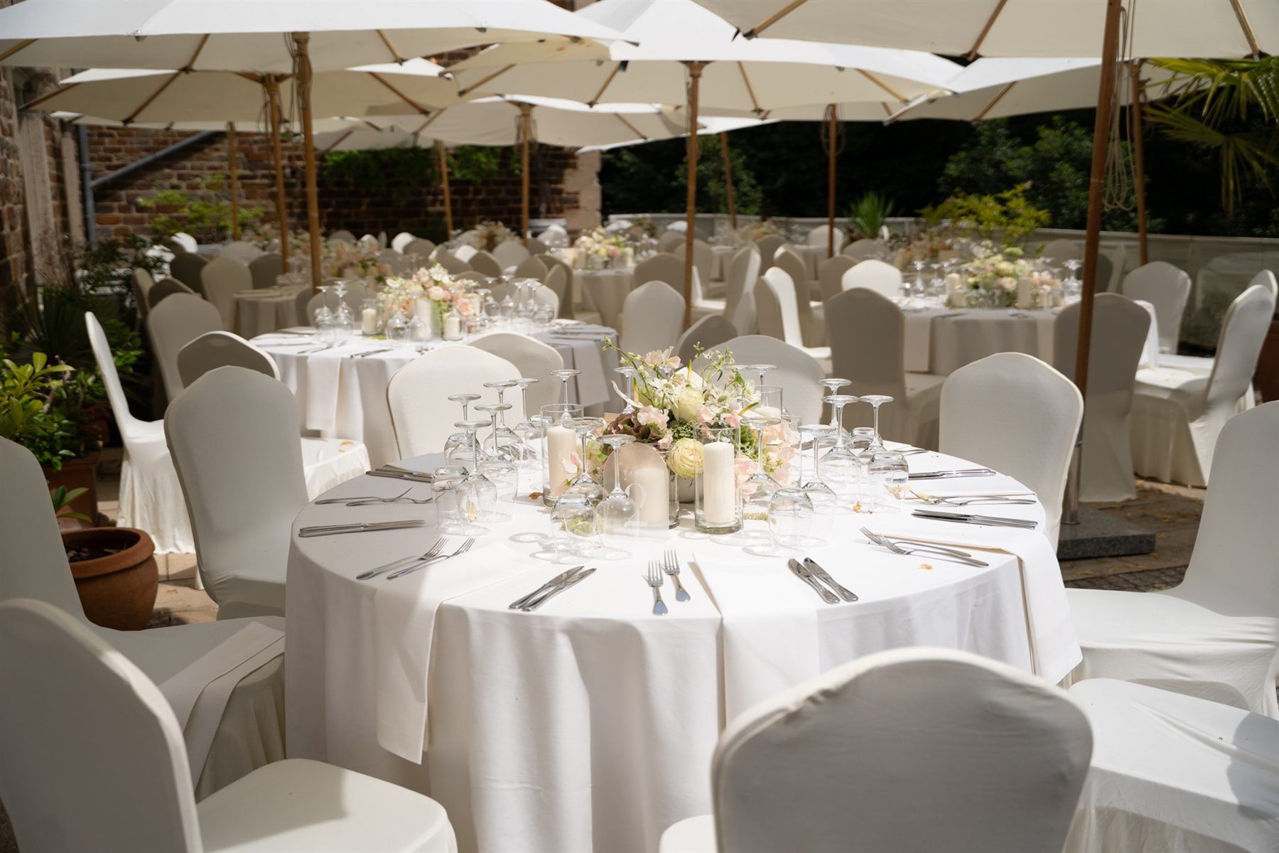Runde Hochzeitstafeln mit weißen Hussen, Kerzen und Blumen unter Sonnenschirmen auf der Schlossterrasse