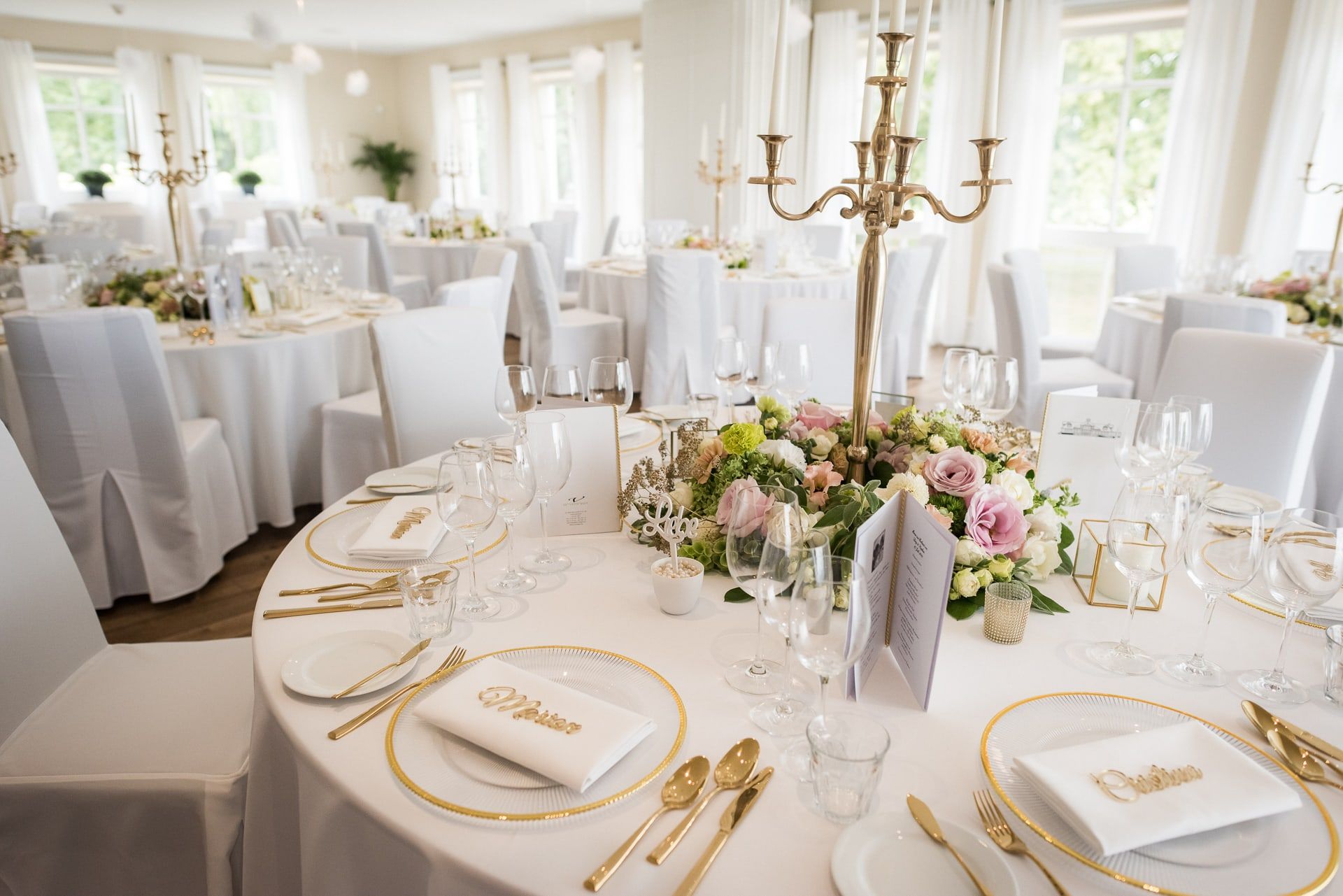 Runde Hochzeitstafel im Festsaal mit Goldbesteck, Kerzenleuchter und Blumen in Pastelltönen