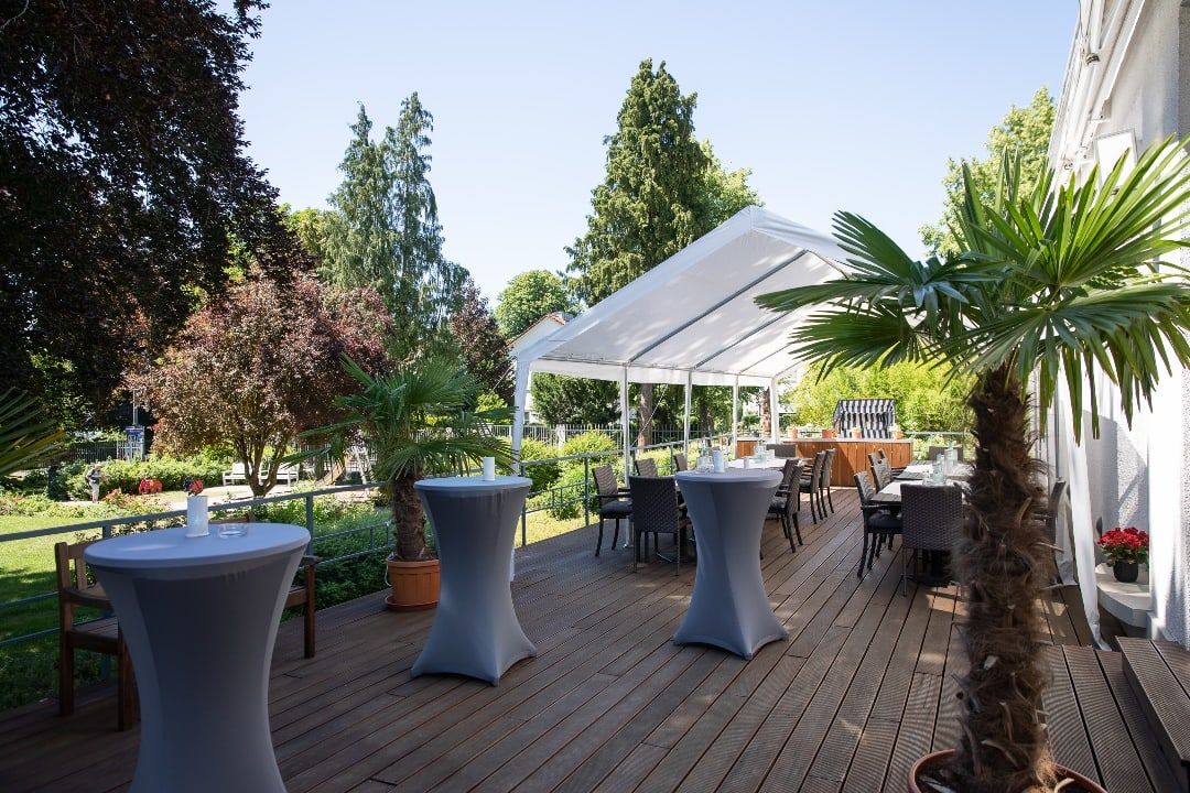 Holzterrasse der Villa Leonhart mit Stehtischen, Palmen und einem weißen Pavillon am Rand des Parks in Königswinter