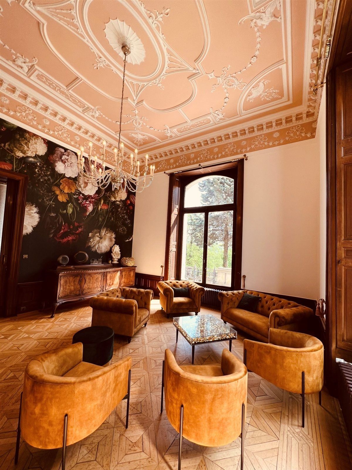 Die Bibliothek-Lounge mit Chesterfield-Sesseln, Stuckdecke, Kronleuchter und hohem Fenster zum Park