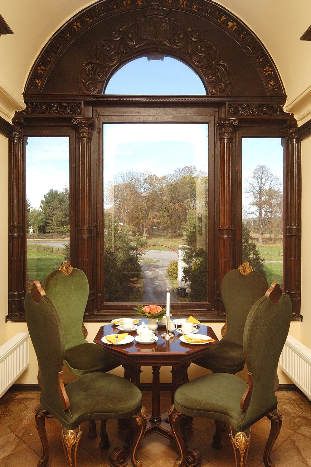 Gedeckter Kaffeetisch mit grünen Samtstühlen vor einem geschnitzten Rundbogenfenster mit Blick auf die Allee von Schloss Wachendorf