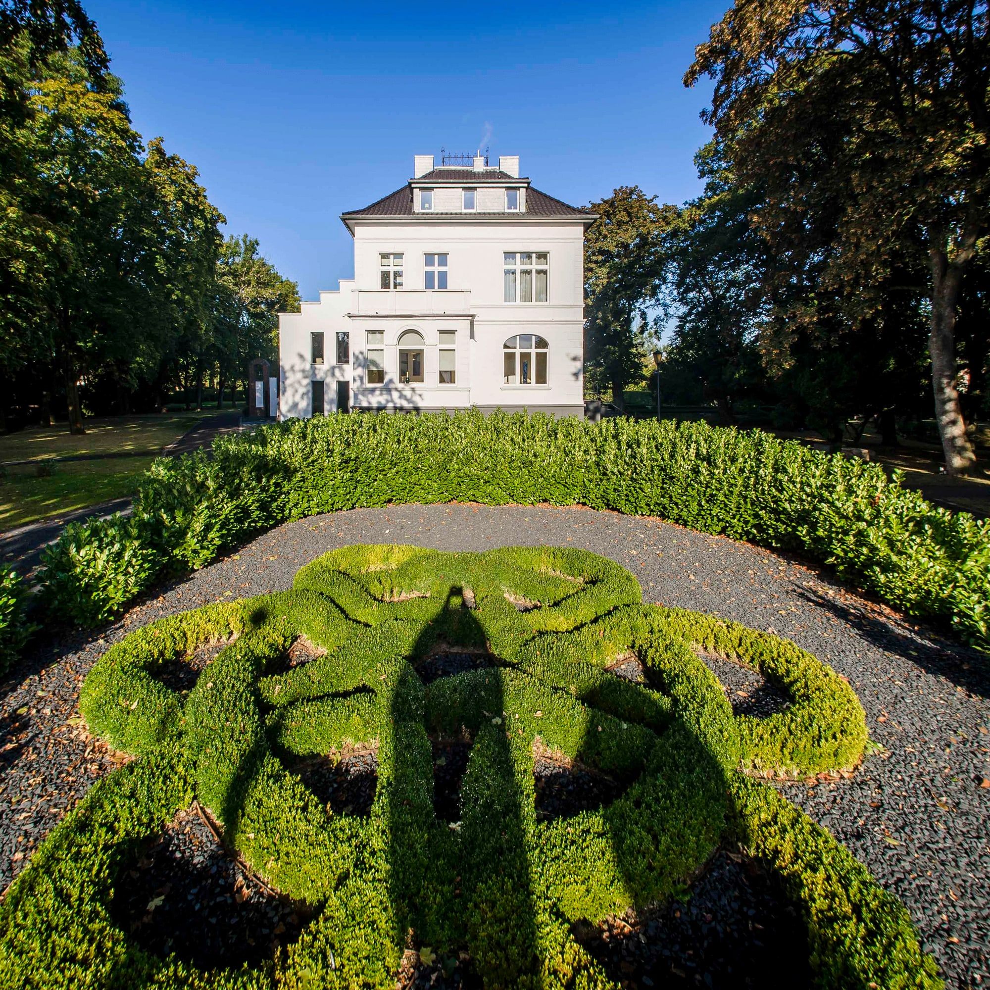 Weiße Villa des Andreashauses in Niederzier hinter einem kunstvoll geschnittenen Buchsornament, eingerahmt von hohen Bäumen