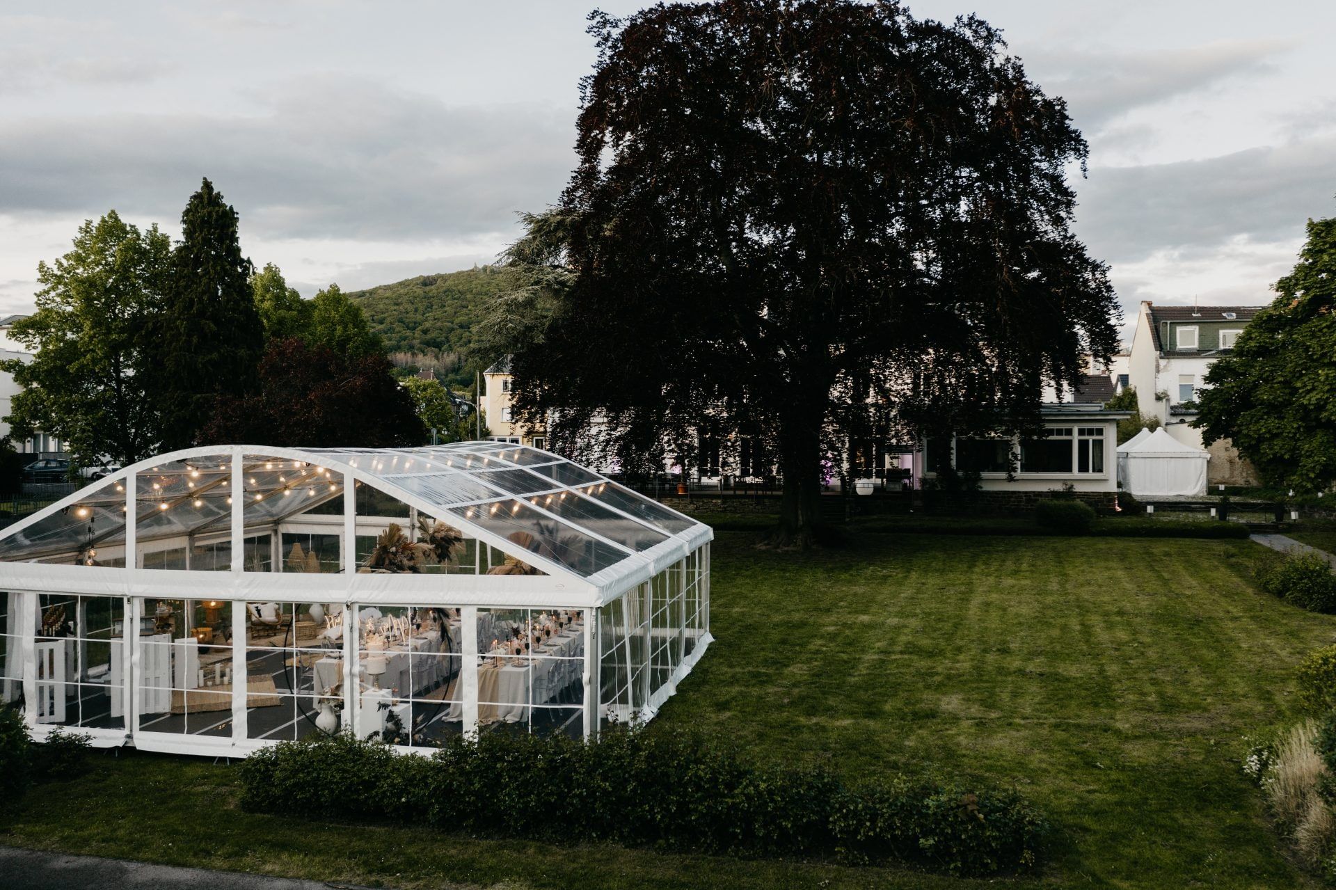 Gläsernes Festzelt mit Lichterketten und eingedeckter Tafel auf dem Rasen im Park der Villa Leonhart unter einer großen Blutbuche