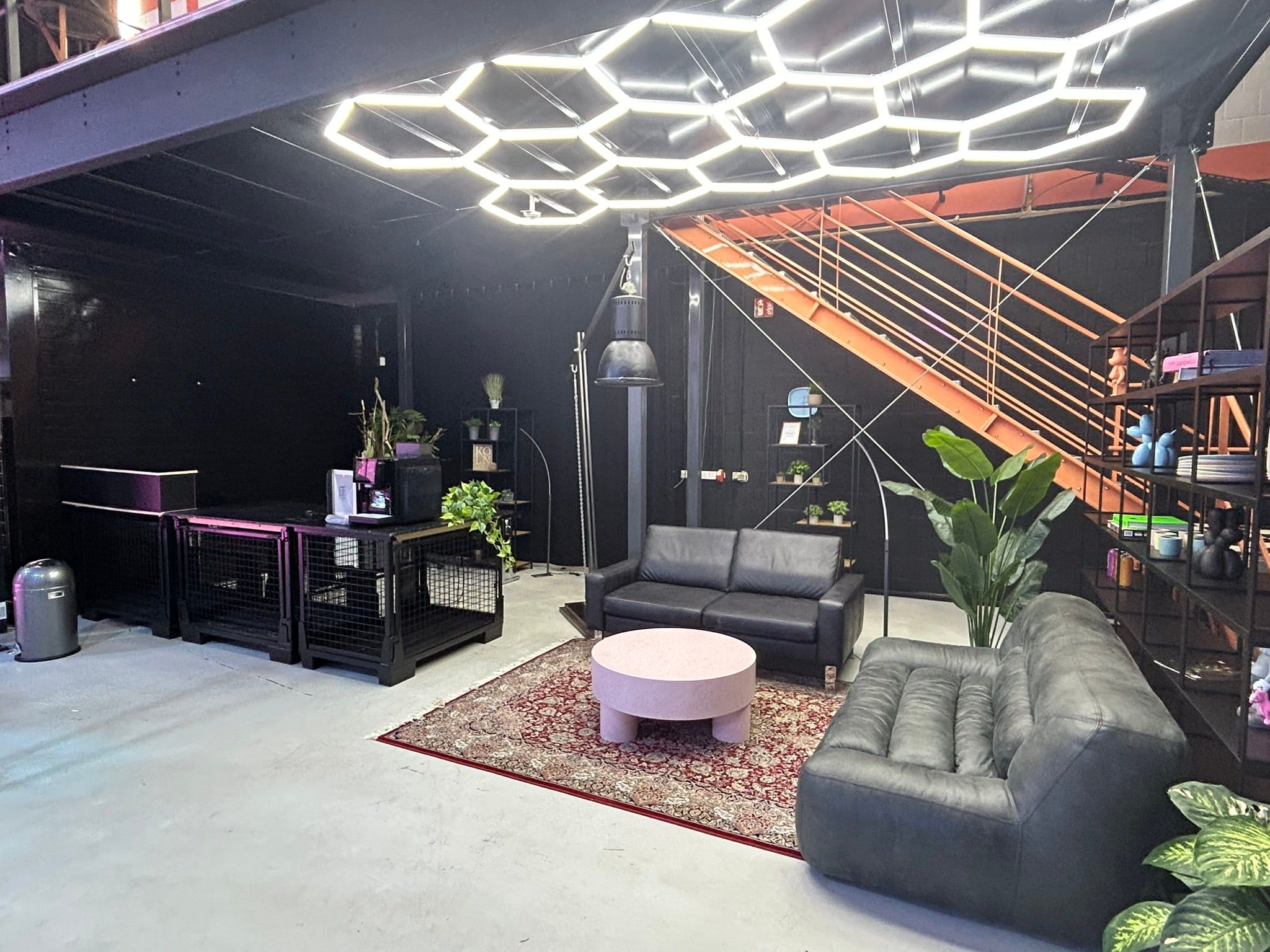 Lounge im Bauwerk Studio unter einer Wabenleuchtdecke, mit orangefarbener Stahltreppe, dunklen Ledersofas und Perserteppich