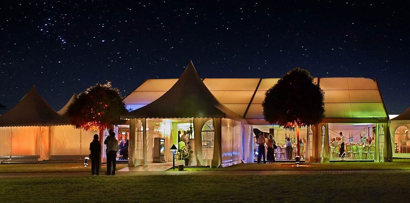Festzeltanlage bei Nacht unter Sternenhimmel, innen warm beleuchtet, davor Bäume und Gäste auf der Wiese