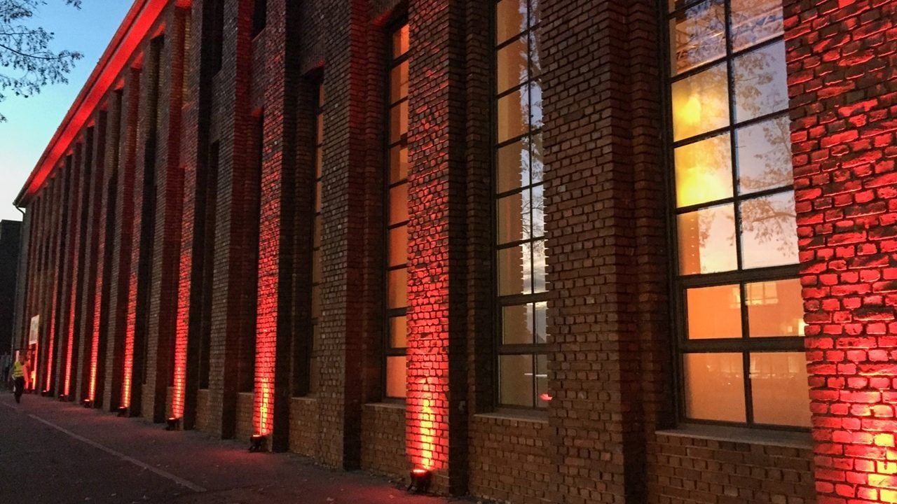 Die Backsteinfassade des Bauwerks in Köln-Kalk am Abend, rot angestrahlt, mit warm leuchtenden Fenstern