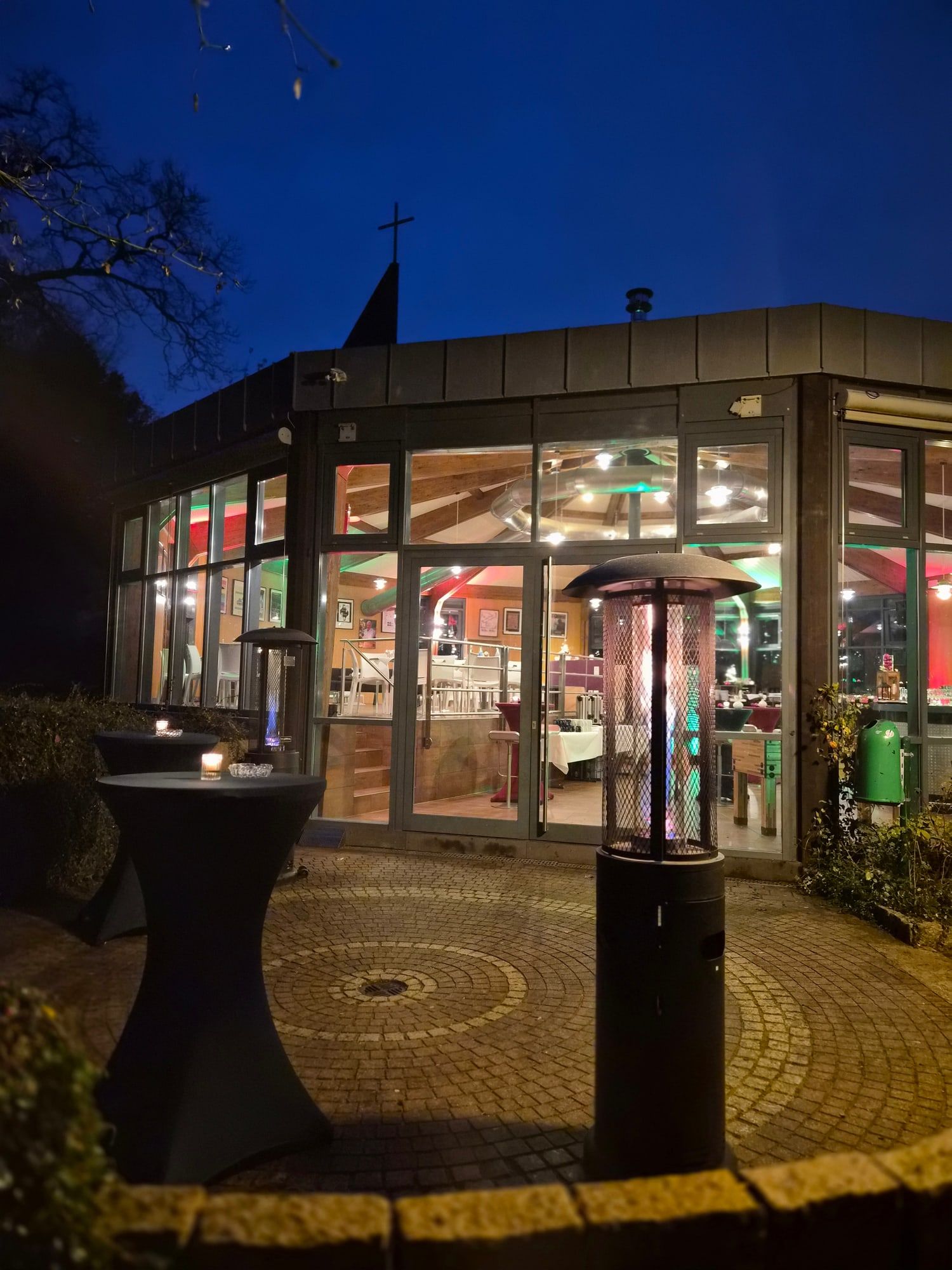 Die Rotunde bei Nacht mit hell erleuchteten Fenstern, gepflastertem Vorplatz, Stehtisch und Heizstrahler