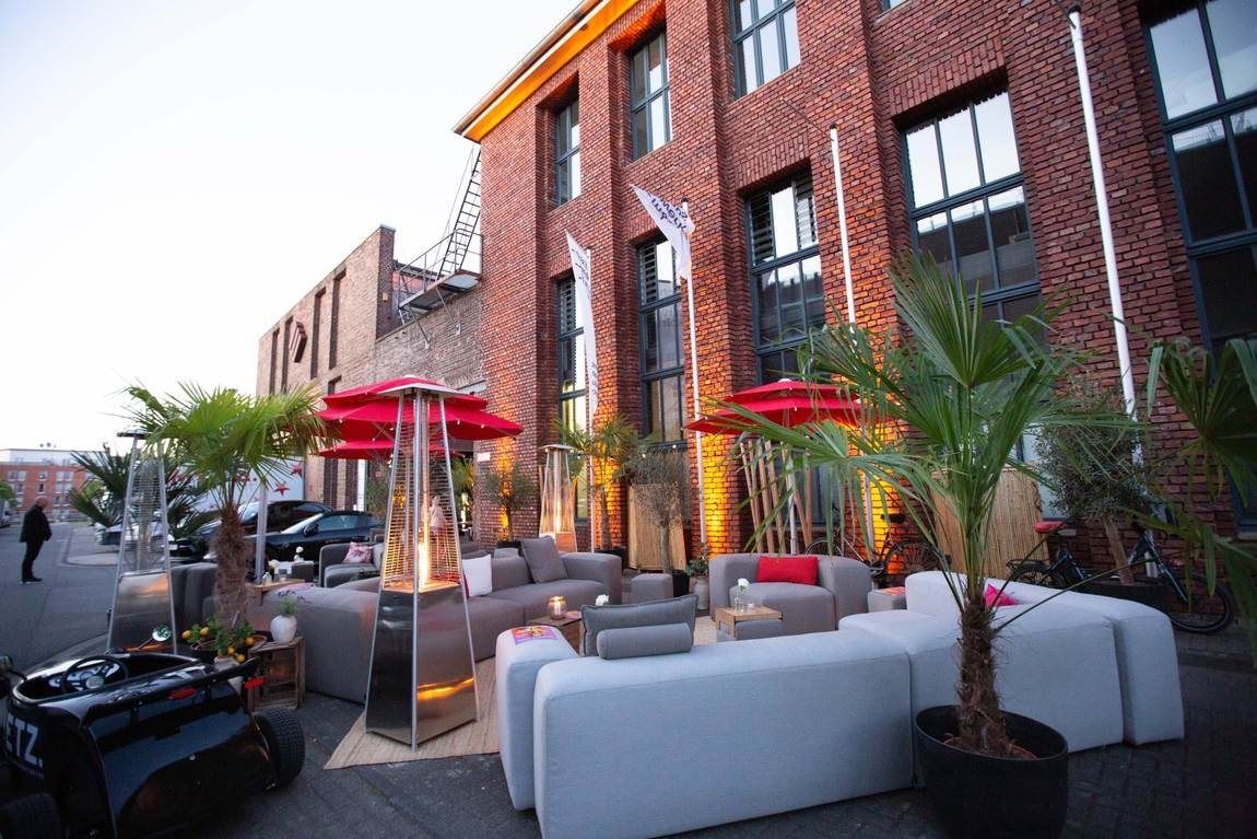 Außenbereich des Bauwerks in Köln am Abend, weiße Lounge-Sofas, Palmen und Heizstrahler vor der Backsteinfassade