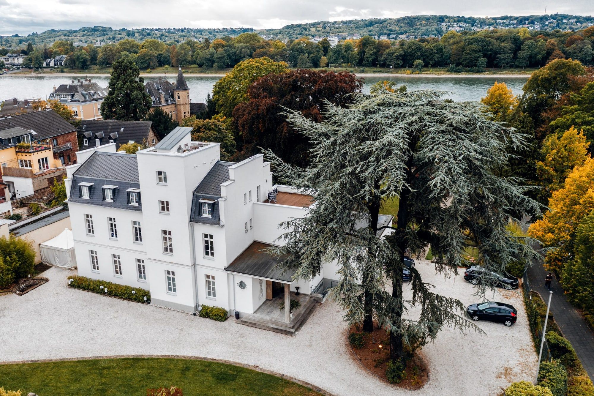 Die weiße Villa Leonhart in Königswinter aus der Luft, mit Schieferdach, alter Zeder und Kiesvorplatz, dahinter der Rhein und das gegenüberliegende Bonner Ufer